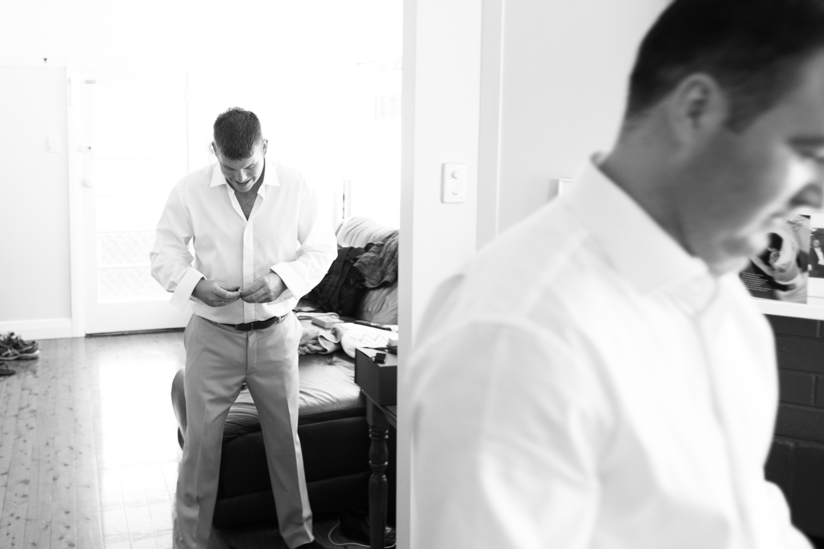 Black and white shot of the groom buttoning up his white shirt while his groomsman walks past in the foreground Caves Beach wedding photography