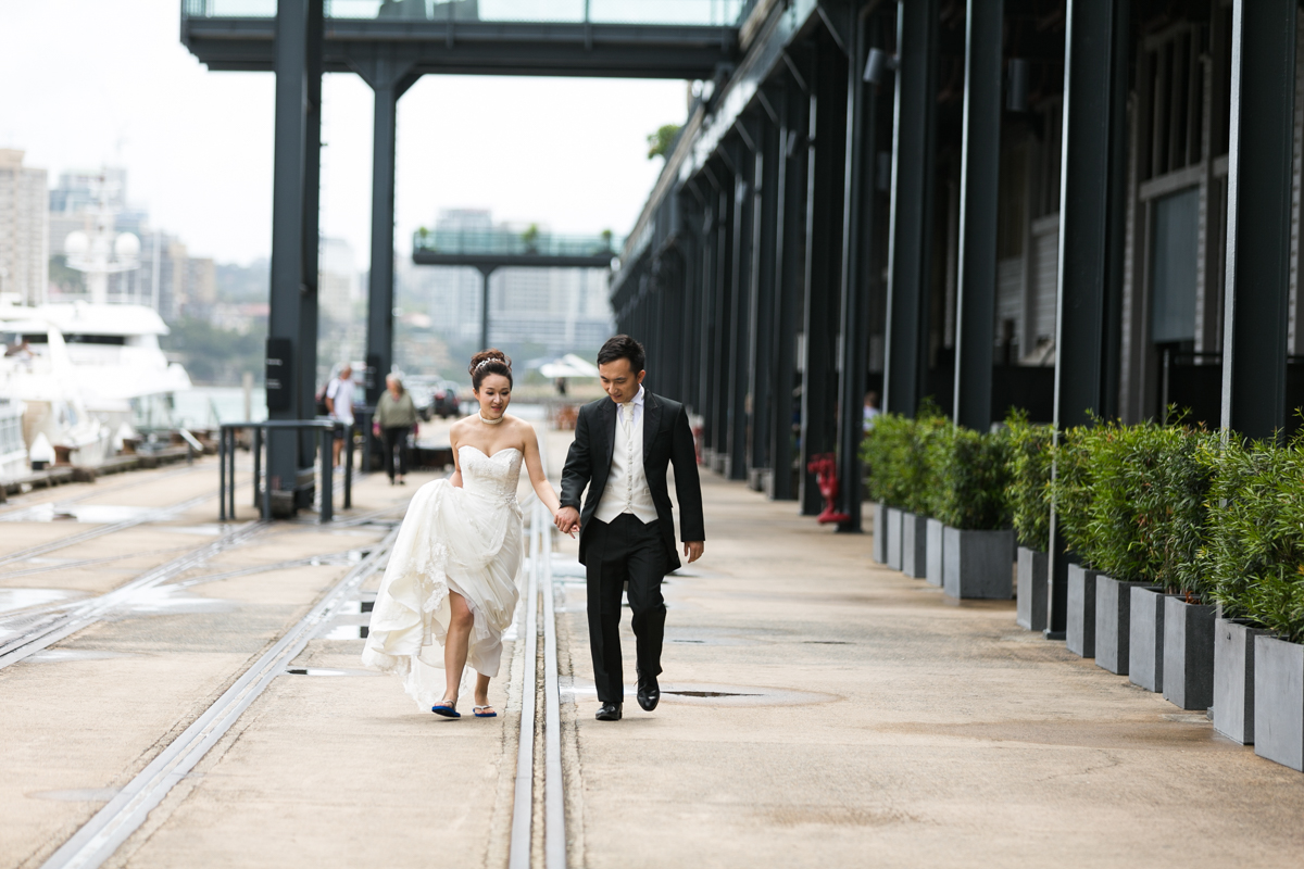 Candid shot of the bride and groom walking along hand in hand at The Rocks in Sydney while she holds her dress up out of the puddles on the ground Sydney wedding photographer