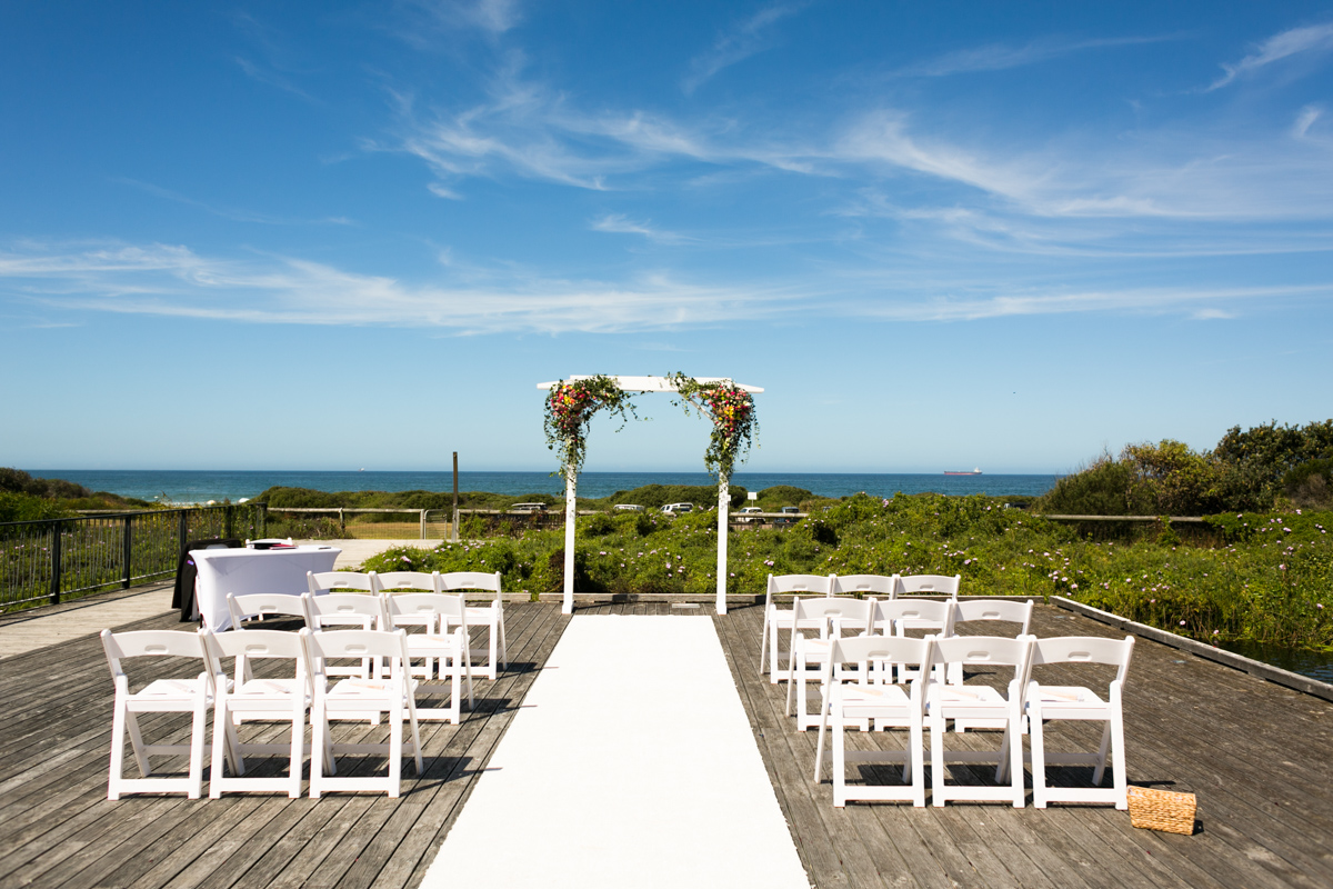 Landscape shot of the wedding ceremony location on a rustic deck overlooking the beach with a white archway decorated with multicoloured flowers and greenery with white deck chairs and a white aisle runner Caves Beach wedding photography