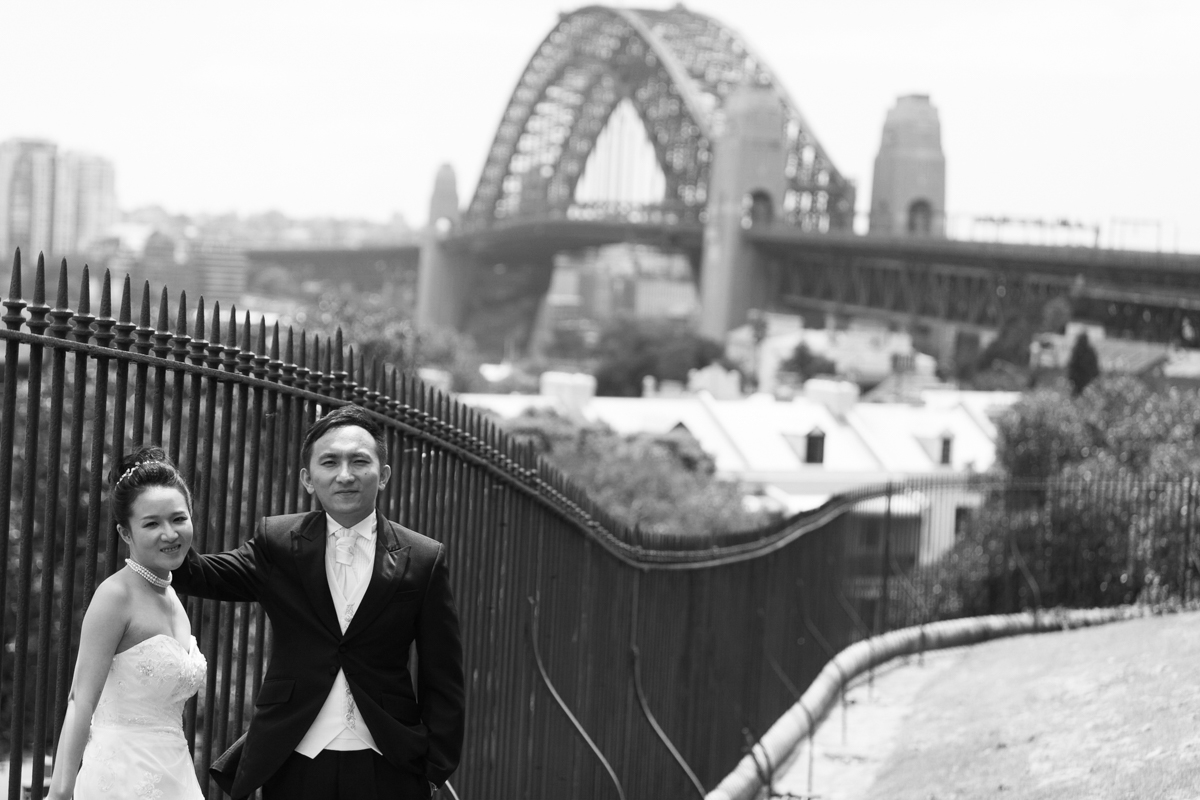 Black and white shot of the bride and groom leaning against a wrought iron fence with the Sydney Harbour Bridge in the background Sydney wedding photographer