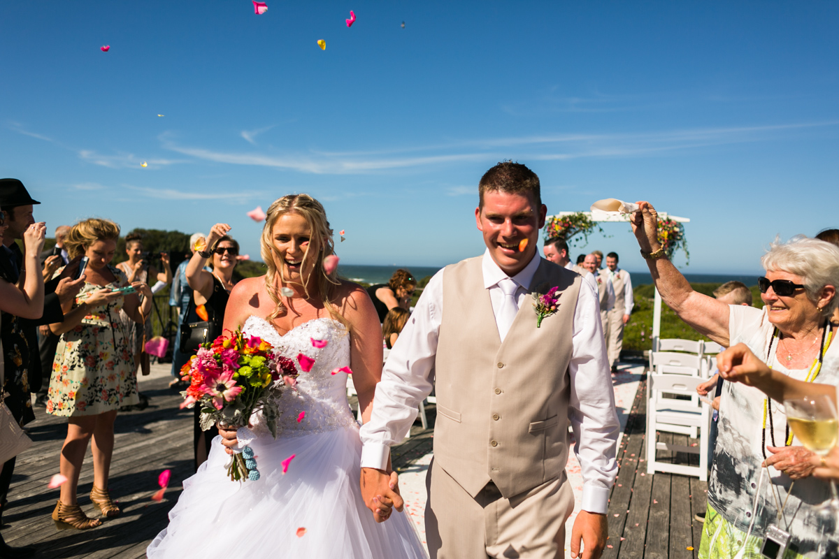 Candid shot of the bride and groom walking back down the aisle while their wedding guests throw rose petals over them Caves Beach wedding photography