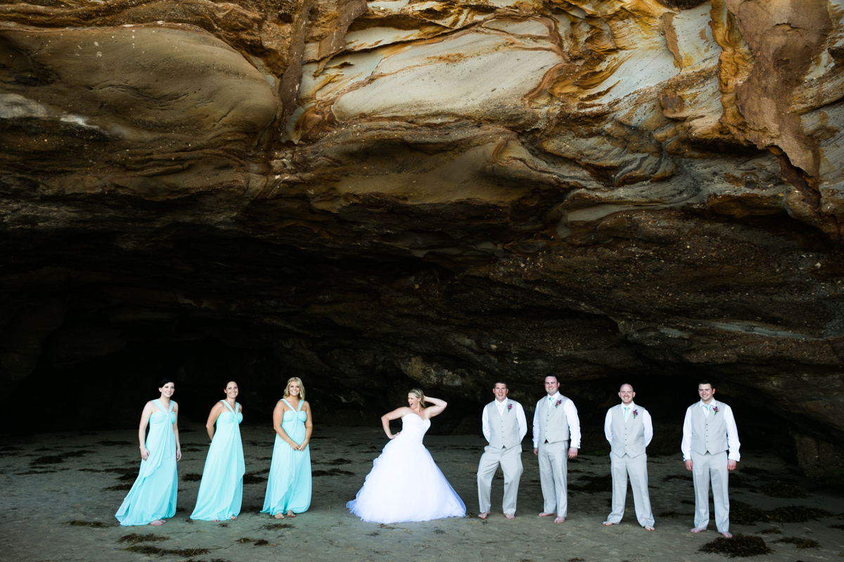 Candid shot of the bride striking a pose flanked by her groom and bridal party standing in the mouth of a natural rock beach cave Caves Beach wedding photography