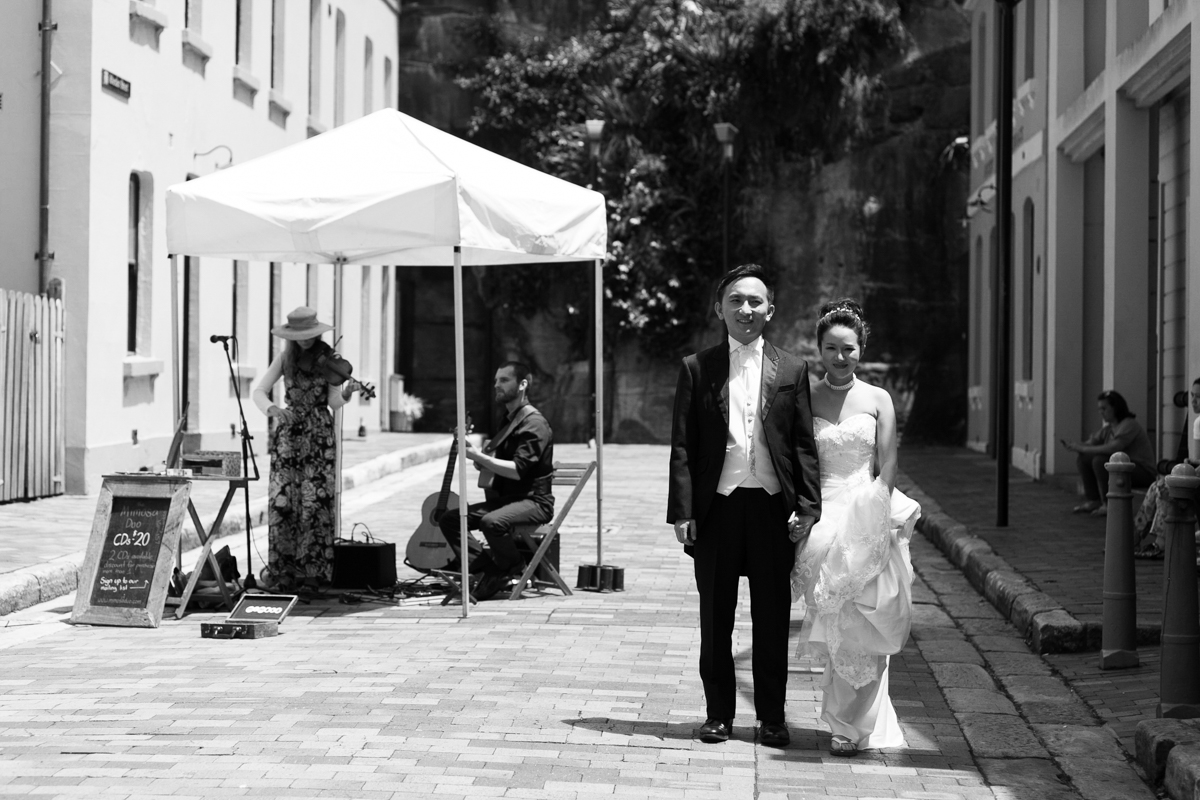Black and white shot of the bride and groom stand holding hands in a cobblestoned alley while a busker plays guitar behind Sydney wedding photographer