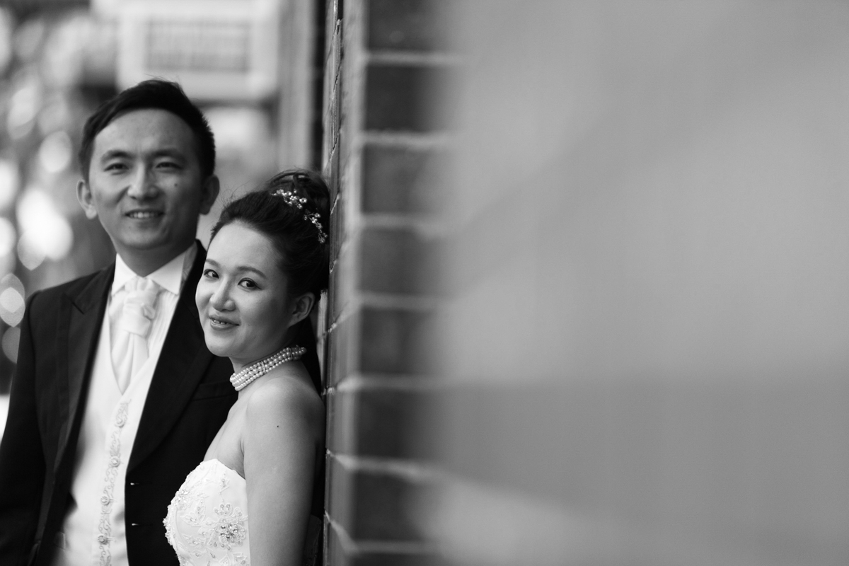 Black and white artistic shot looking down a brick wall toward the bride and groom who are leaning against the wall looking into the camera Sydney wedding photographer