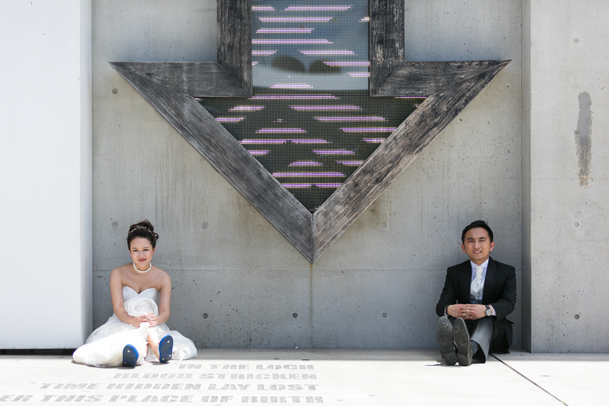 Artistic shot of the bride and groom seated in front of a cement wall under a lit up wooden arrow art installation at the Museum of Contemporary Art Sydney wedding photographer