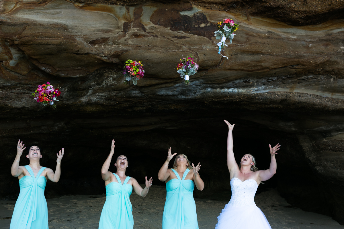 The bride and bridesmaids stand in a line in the mouth of a natural rock beach cave and throw their multicoloured flower bouquets in the air Caves Beach wedding photography