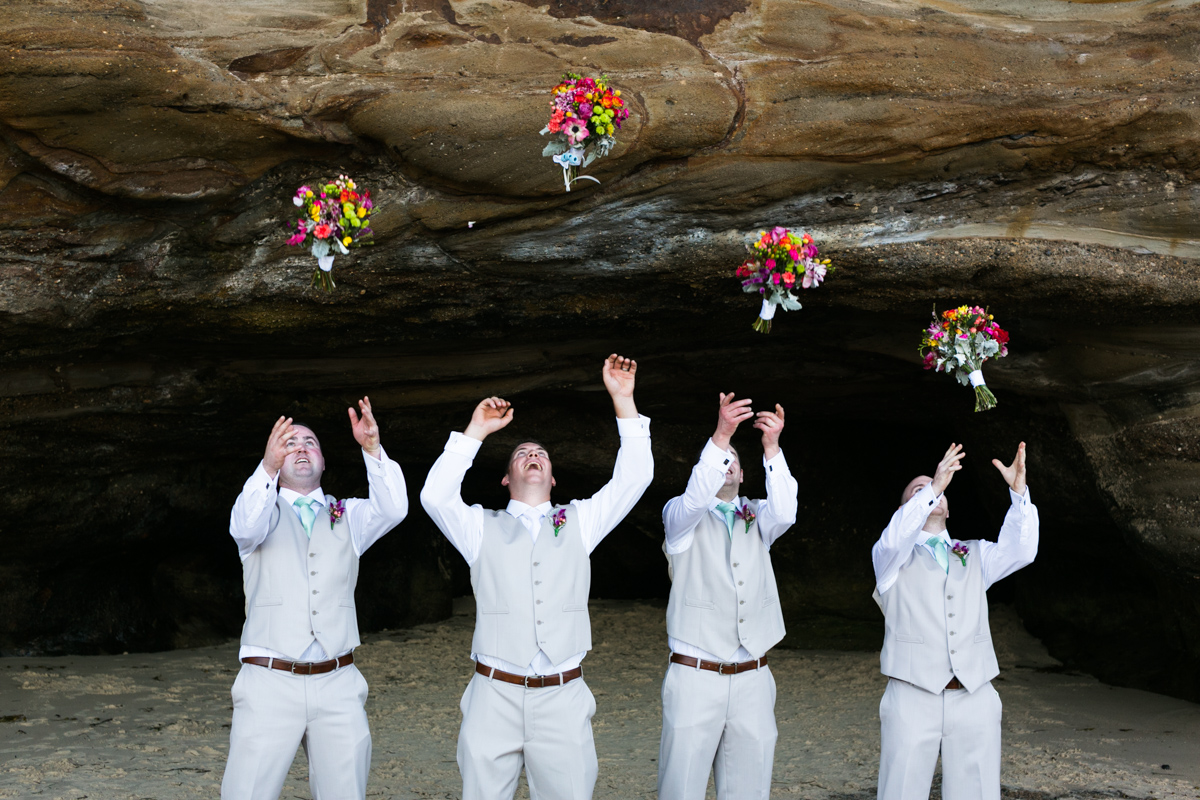 The groom and his groomsmen stand in a line in the mouth of a natural rock beach cave throwing the bridesmaid's multicoloured bouquets in the air Caves Beach wedding photography