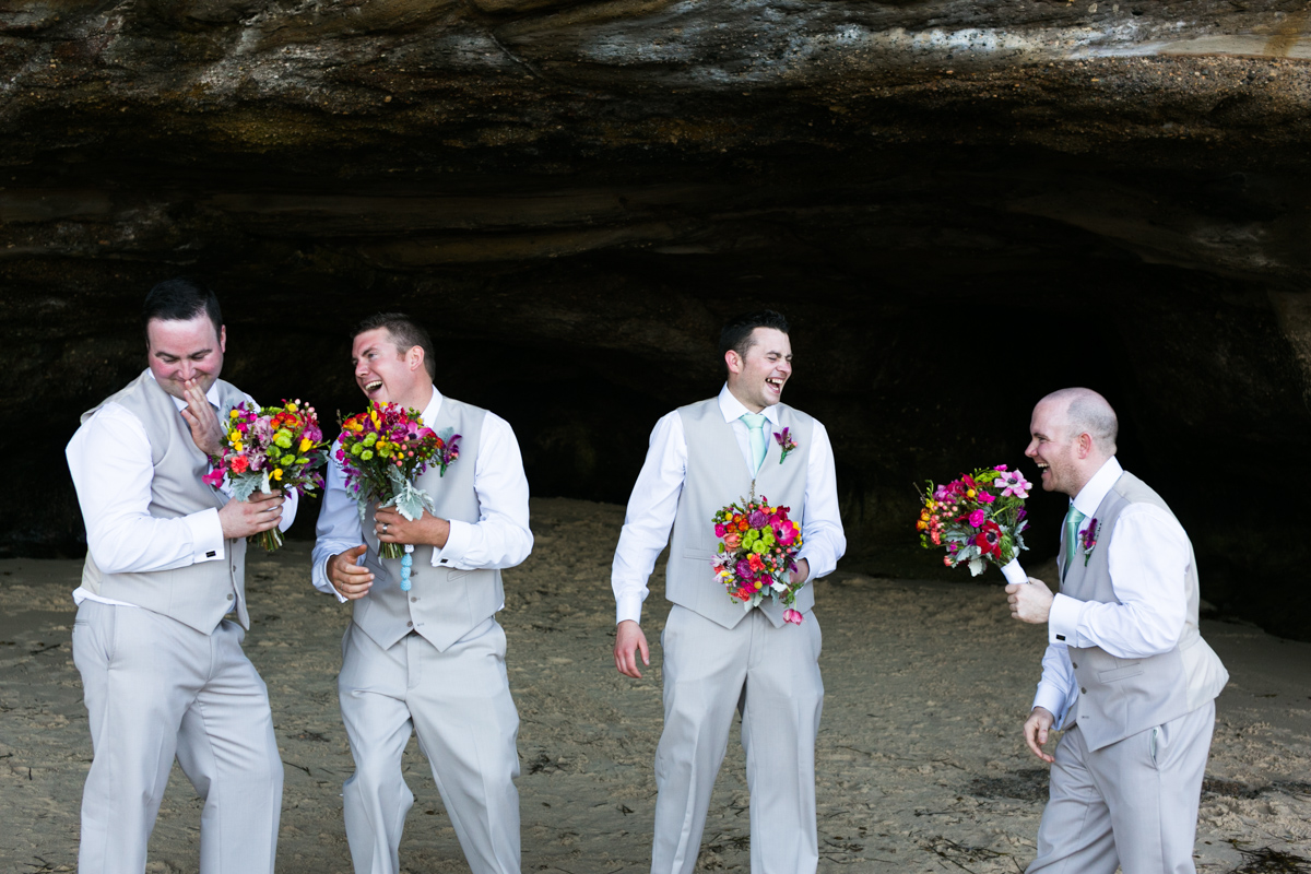 Candid shot of the groom and groomsmen being silly with the multicoloured bridal bouquets in the mouth of a beach cave Caves Beach wedding photography