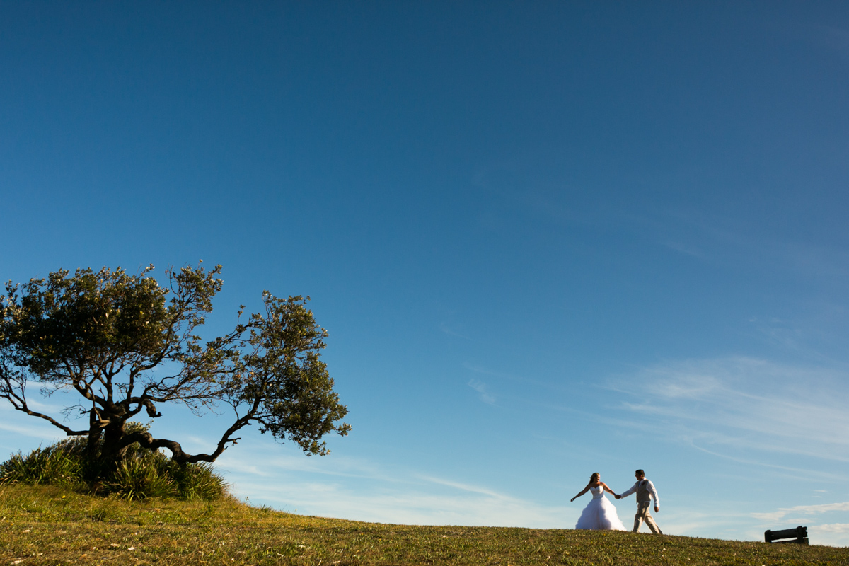 Landscape shot of the bride and groom holding hands walking up a green hill with clear blue skies behind Caves Beach wedding photography