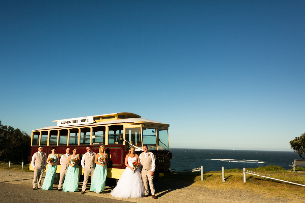 Landscape shot of the bride and groom standing with their bridal party in front of Newcastle's Famous Tram with the ocean and blue sky in the background Caves Beach wedding photography
