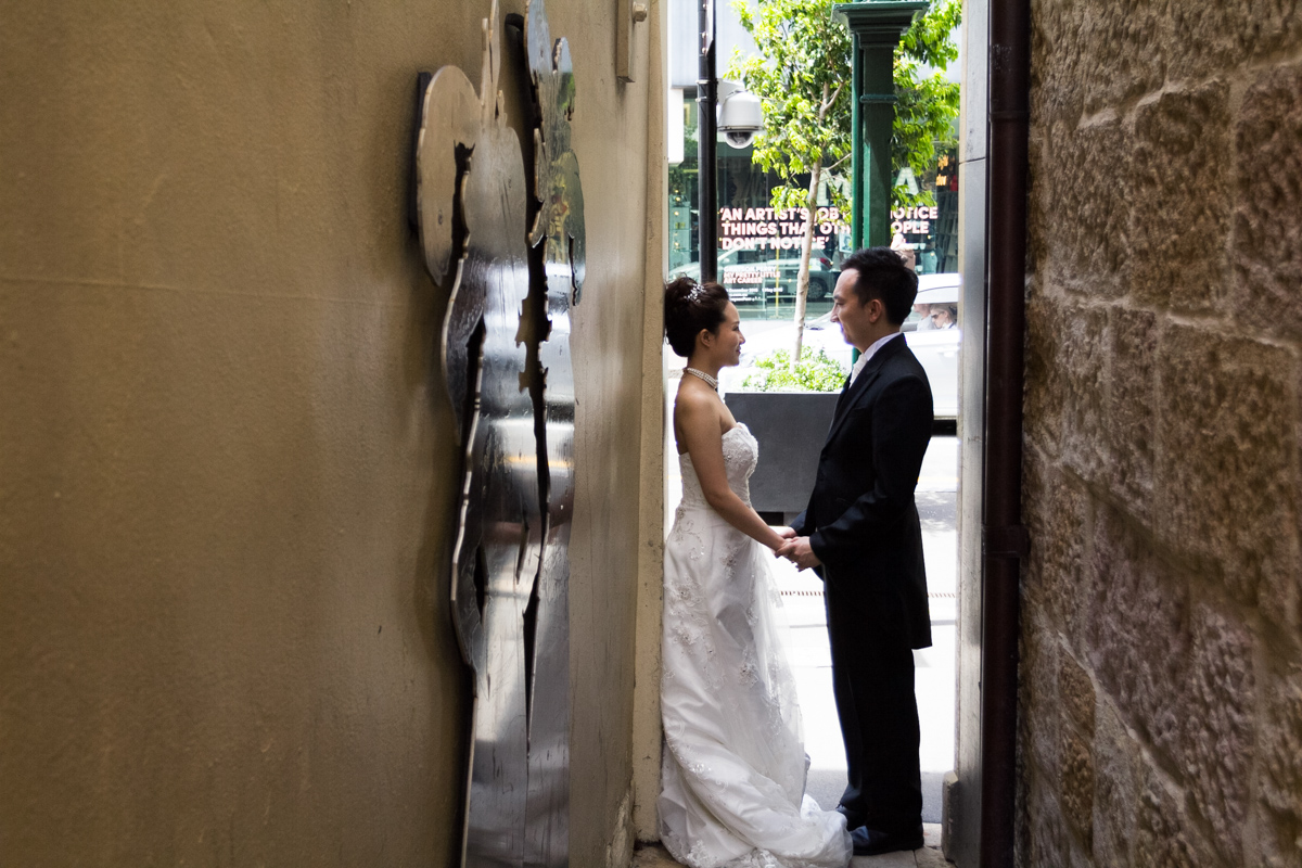 The bride and groom stand facing each other holding hands shot down the length of a brick and cement alleyway with a silver art installation on one wall Sydney wedding photographer